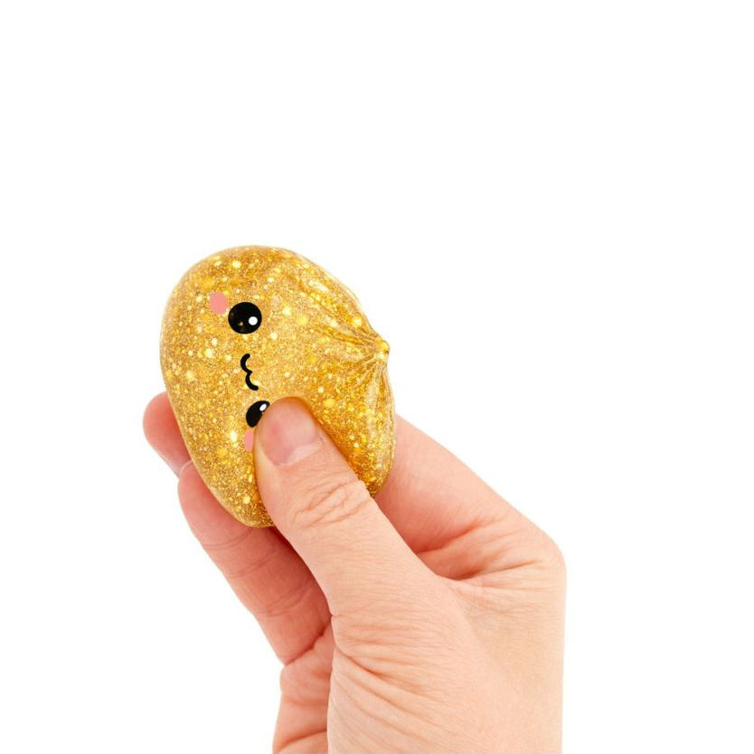 Hand gently squeezing a small gold glitter dumpling‑shaped squishy toy with a smiling cartoon face, shown against a white background.