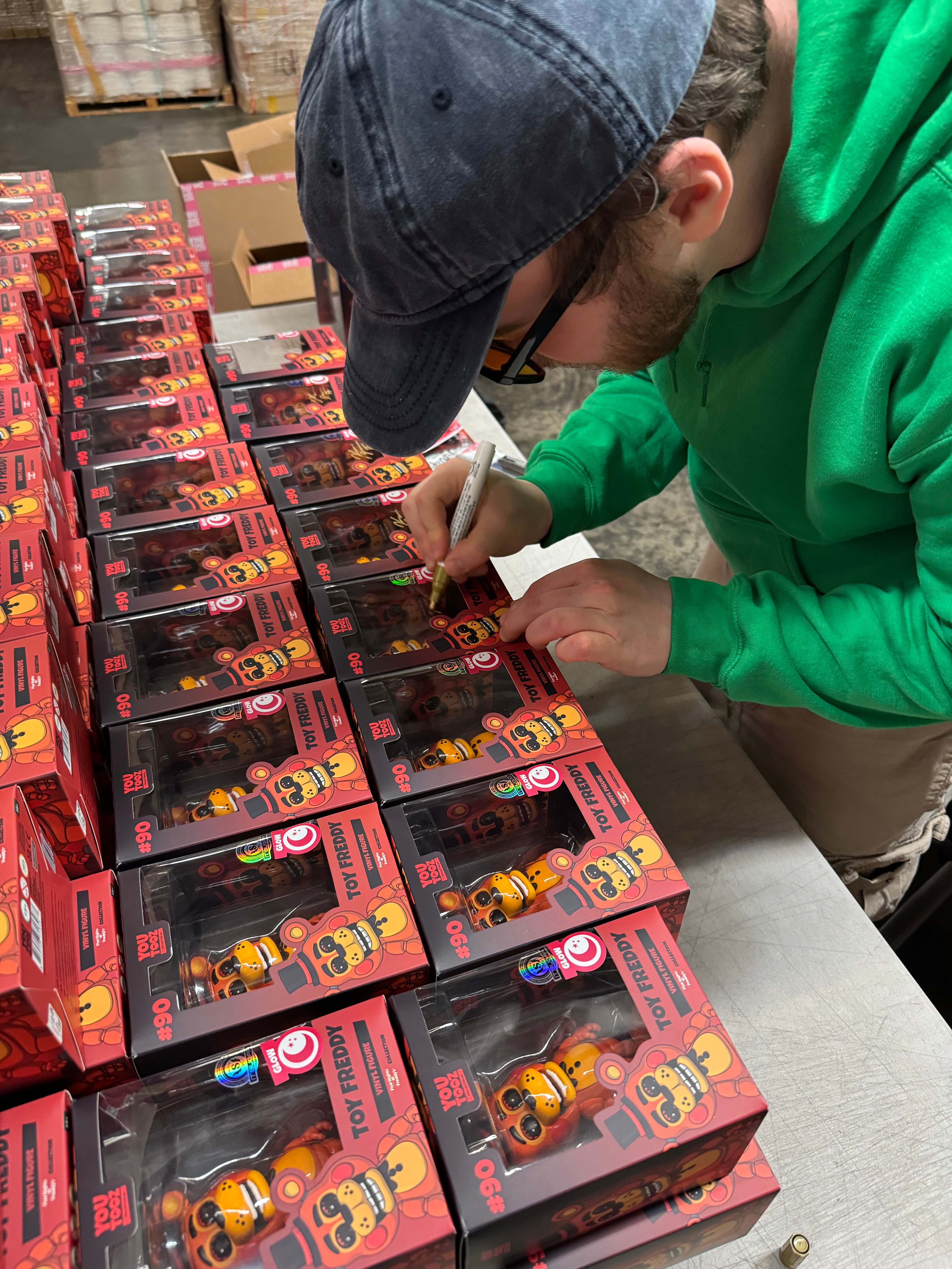 Kellen Goff in a green hoodie and blue cap signs collectible toy vinyl boxes, arranged in rows on a table in a warehouse or studio setting.