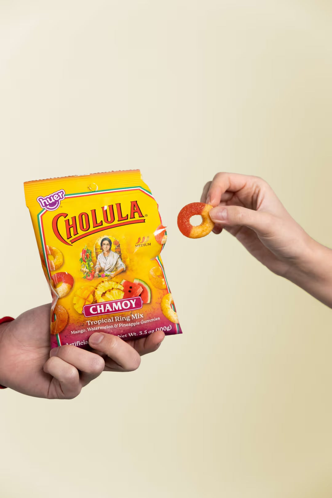 A person holds a yellow bag of Cholula Chamoy Tropical gummy ring mix in one hand while another hand holds up a round ring-shaped piece of the chew, against a plain light beige background.