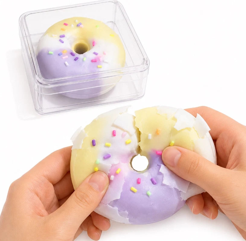 A squishy donut-shaped toy with a pastel yellow and lavender purple color scheme and multicolored sprinkles, shown being held open by two hands. A clear plastic storage case containing a similar closed donut is visible in the background on a white surface.