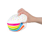 A hand holding a white cake-shaped squishy toy with colorful rainbow stripes and small multicolored dots, shown against a plain white background.