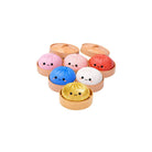 Seven dumpling‑shaped squishy toys in different colors—pink, light pink, red, blue, white, and gold—each with a small smiling cartoon face, displayed individually inside round tan containers with matching lids on a white background.