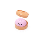 Open round tan container with the lid hovering above, holding a pink dumpling‑shaped squishy toy with a small smiling cartoon face, shown on a white background.