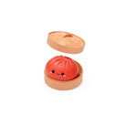 Open round tan container with the lid slightly raised, holding a red dumpling‑shaped squishy toy with a small smiling cartoon face, shown on a white background.