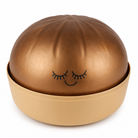 A metallic bronze squishy dumpling with a matching beige base, featuring a simple black line drawing of a sleeping face on the front, set against a plain white background.