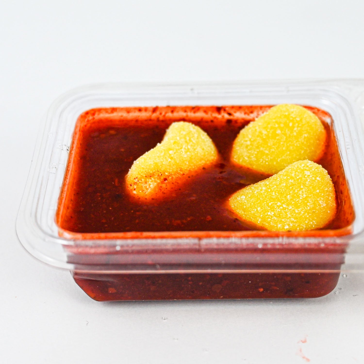 A clear plastic clamshell container holding a chamoy dipping sauce with three yellow, sugar-coated heart-shaped candies on top, photographed on a plain white background.