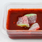 A close-up of a clear plastic container filled with chamoy dip, with three sugar-dusted fruit pieces resting on top.