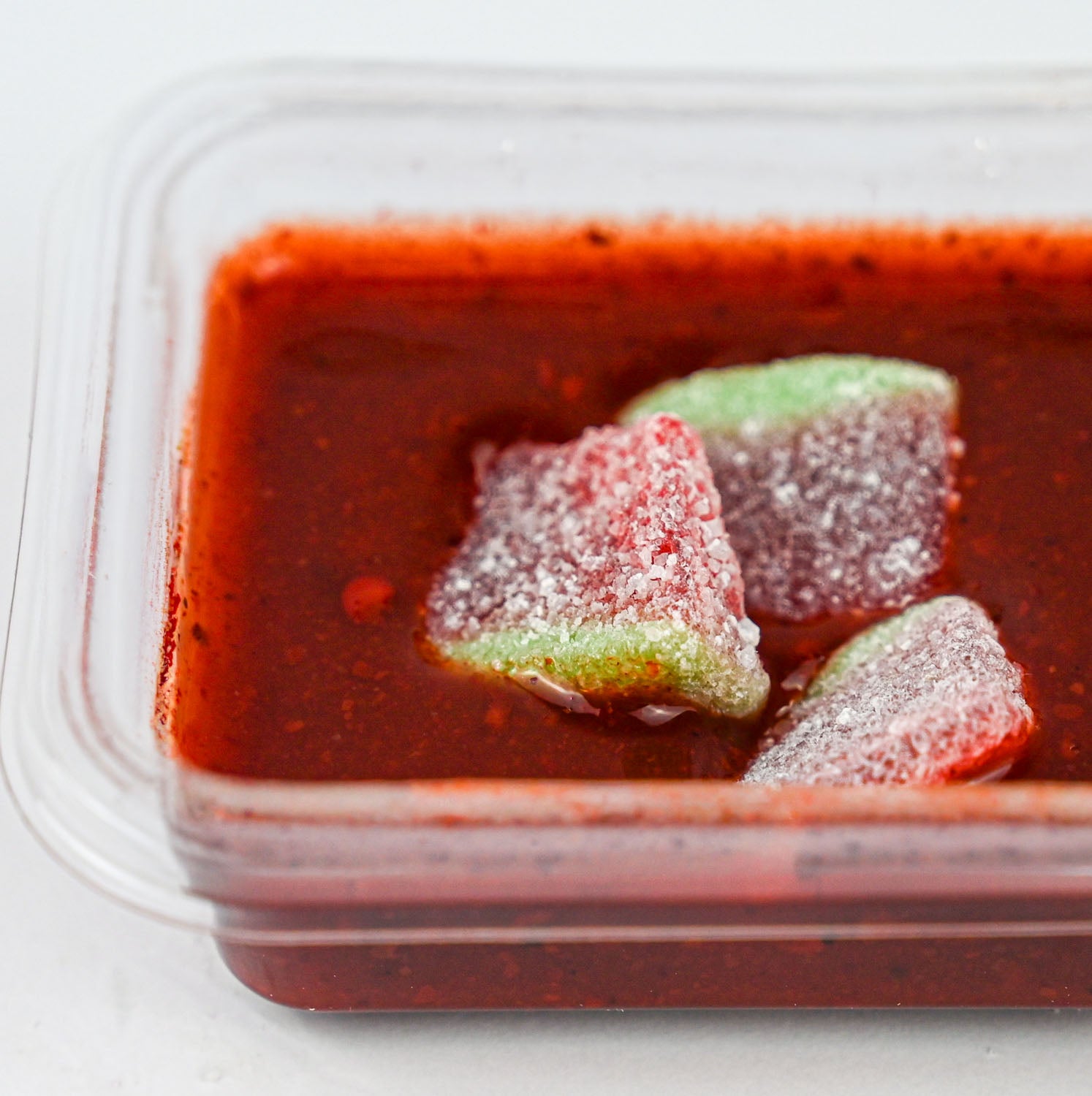 A close-up of a clear plastic container filled with chamoy dip, with three sugar-dusted fruit pieces resting on top.