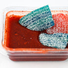 A close-up of a clear plastic container filled with red candy, featuring two large, sugar-coated triangular gummy candies in blue-green and red-orange.