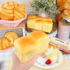 Hand holding a hawaiian bread squishy toy with a background of various bread items and kitchenware.