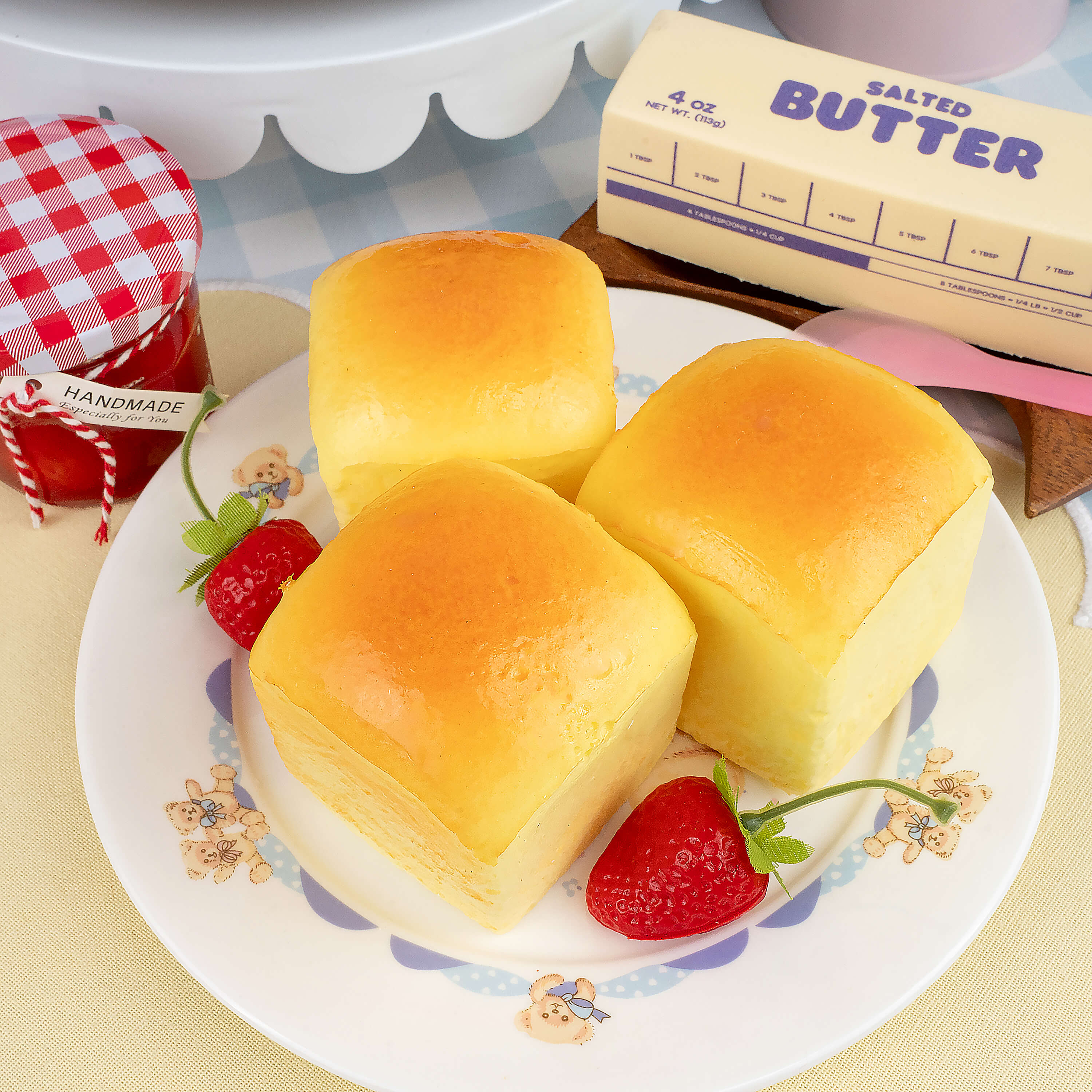 Three Hawaiian bread squishy toys on a decorative plate with strawberries and a stick of salted butter in the background.