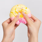 A close-up of two hands holding and crushing a pastel yellow and pink donut squishy toy with a cracked, broken surface and small pink sprinkles, against a light gray background.