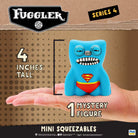 A blue Fuggler Series 4 Mini Squeezables mystery figure with a red and yellow Superman-style chest emblem, shown resting on a person's hand against a brown cardboard-style background with size and branding text.
